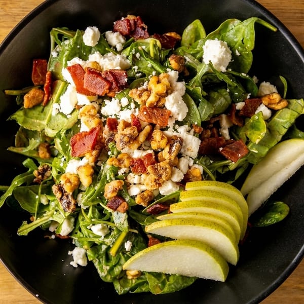 A vibrant salad with spinach, arugula, bacon, walnuts, feta cheese, and sliced pears in a black bowl.