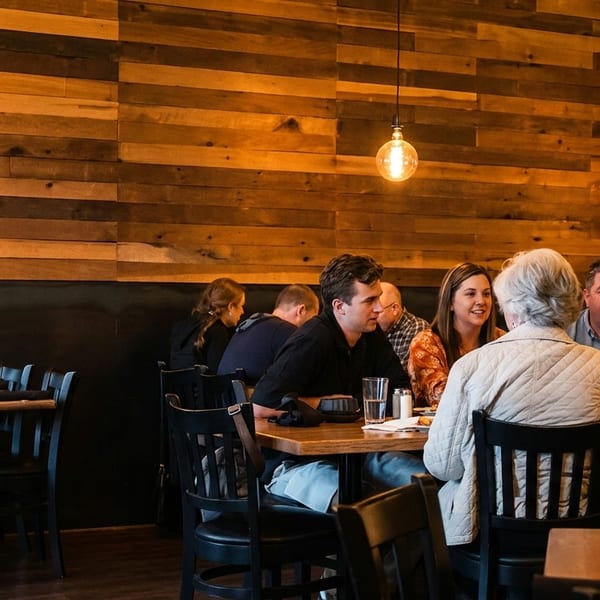 Diners seated at wooden tables in a restaurant with a rustic wood-paneled wall and Edison bulb lighting.