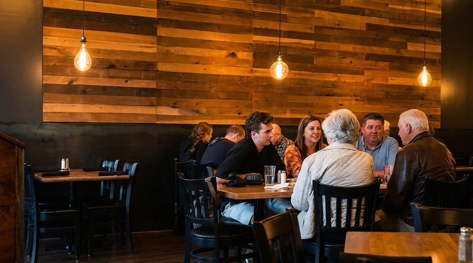 Diners seated at wooden tables in a restaurant with a rustic wood-paneled wall and Edison bulb lighting.