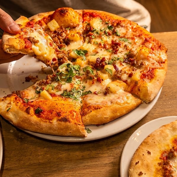 A hand takes a slice of a pineapple and sausage pizza from a white plate on a wooden table.