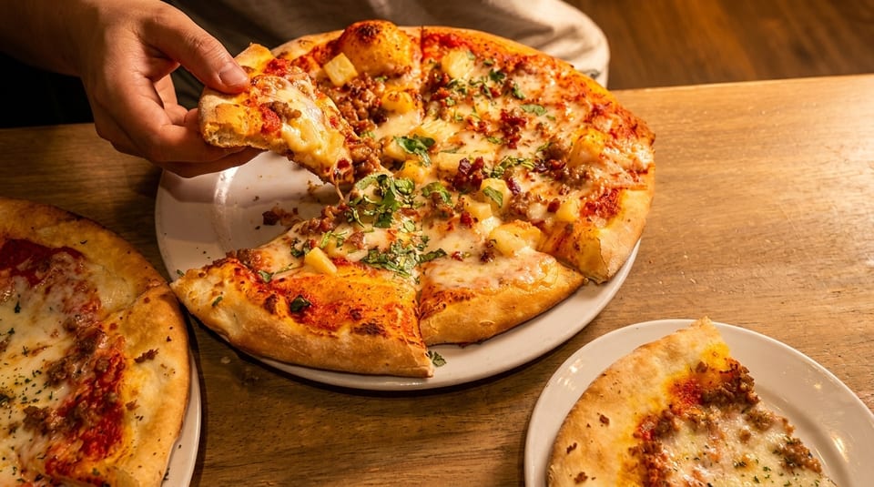 A hand takes a slice of a pineapple and sausage pizza from a white plate on a wooden table.
