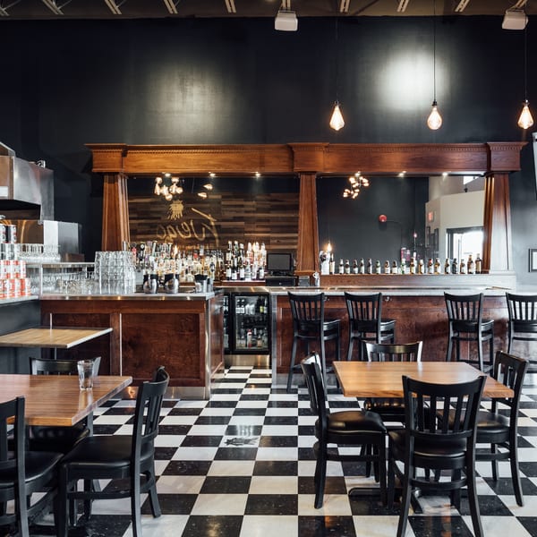 Restaurant interior with black and white checkered floor, wooden tables, black chairs, and a full bar.