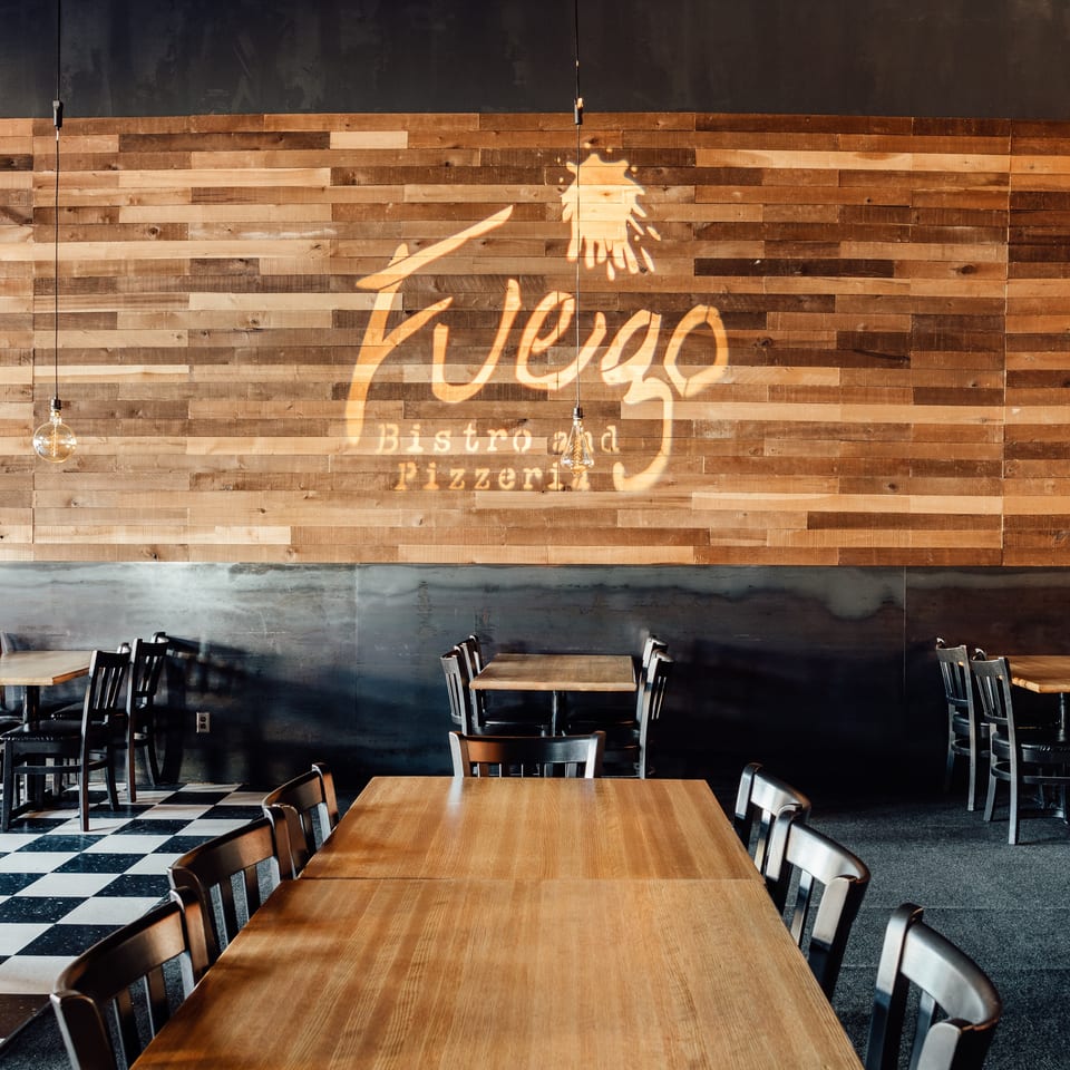 Rustic pizza bistro dining room with wooden tables, black chairs, pendant lights, checkerboard flooring, and a large wall logo.