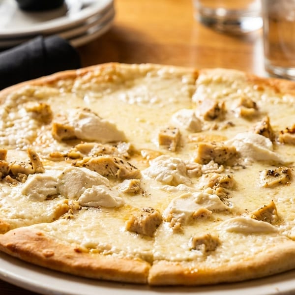 White pizza with chicken and ricotta cheese on a white plate, with a black napkin and glasses in the background.