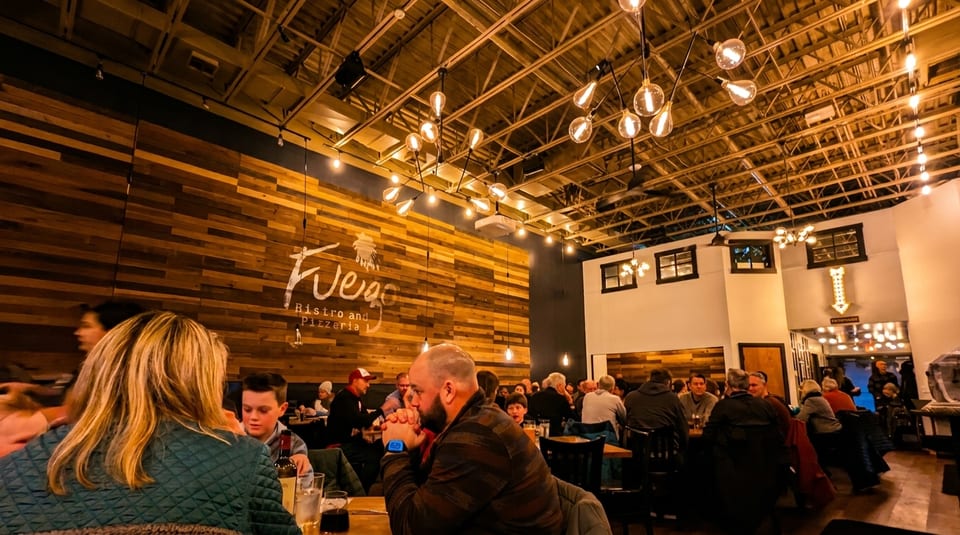 Diners seated at tables in a dimly lit restaurant with a wooden accent wall and industrial lighting.