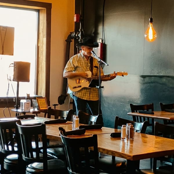 Musician in a hat playing guitar and singing in a restaurant with wooden tables and chairs.