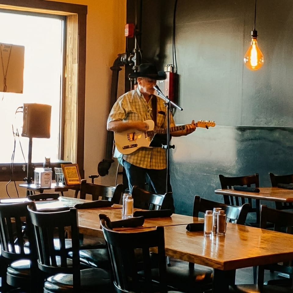 Musician in a hat playing guitar and singing in a restaurant with wooden tables and chairs.