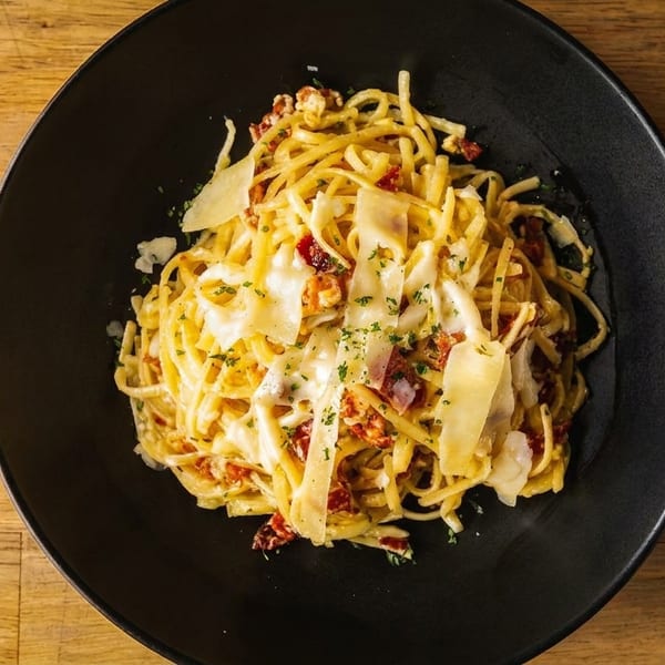 Creamy pasta with sun-dried tomatoes, bacon, and parmesan cheese in a black bowl on a wooden table.