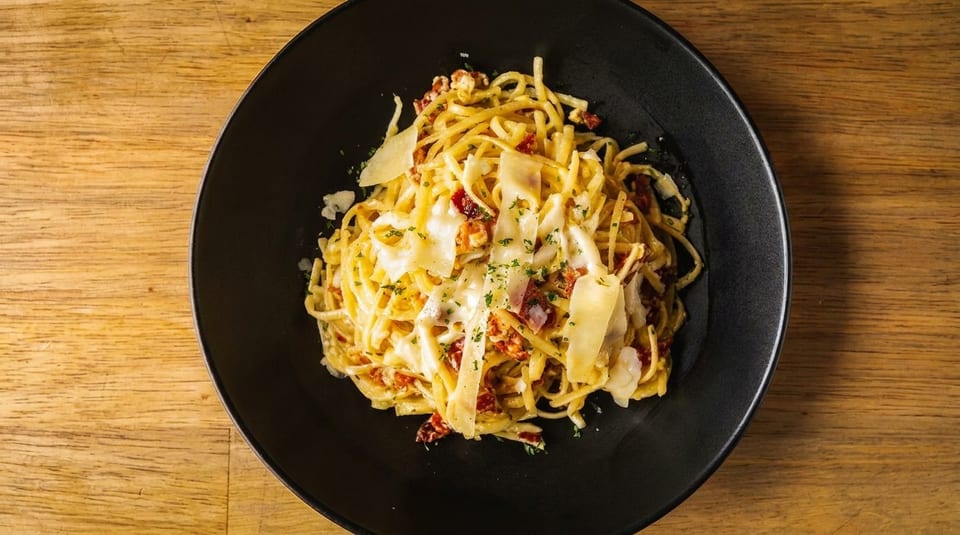 Creamy pasta with sun-dried tomatoes, bacon, and parmesan cheese in a black bowl on a wooden table.