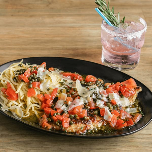 Chicken piccata with pasta, tomatoes, capers, and shaved cheese, alongside a pink cocktail.